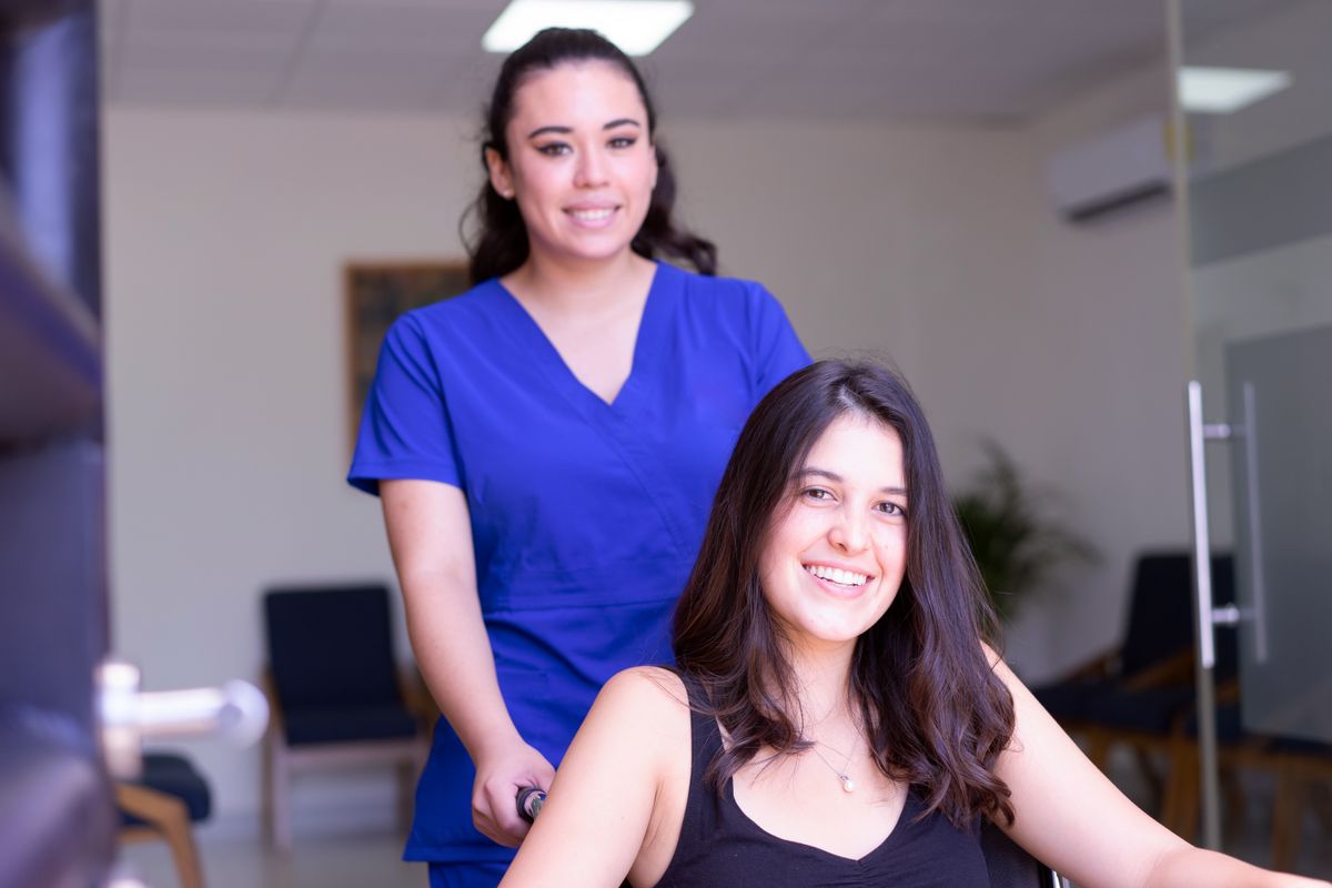 A nurse in blue scrubs assists a smiling patient in a wheelchair indoors